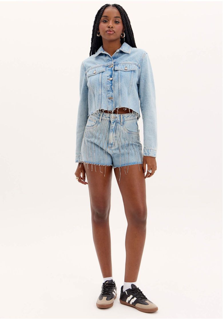 Modelo em pé usando short jeans Grandfather Brilho e jaqueta cropped - vista frontal. Short cintura alta em lavagem azul com franjas de brilho frontais e barra desfiada, look casual glam.