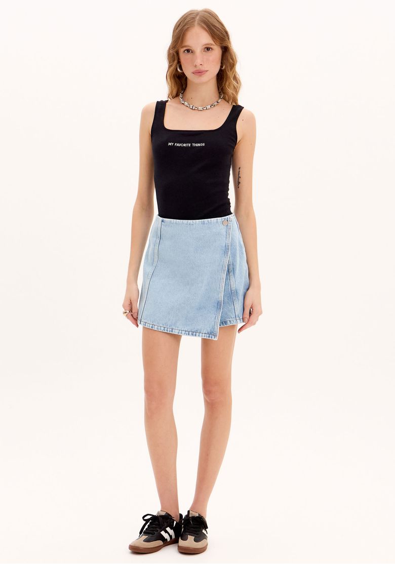 Modelo vestindo um short saia jeans de cintura alta, lavagem clara, visualizado de frente, combinado com uma regata preta e tênis - short saia denim feminino.