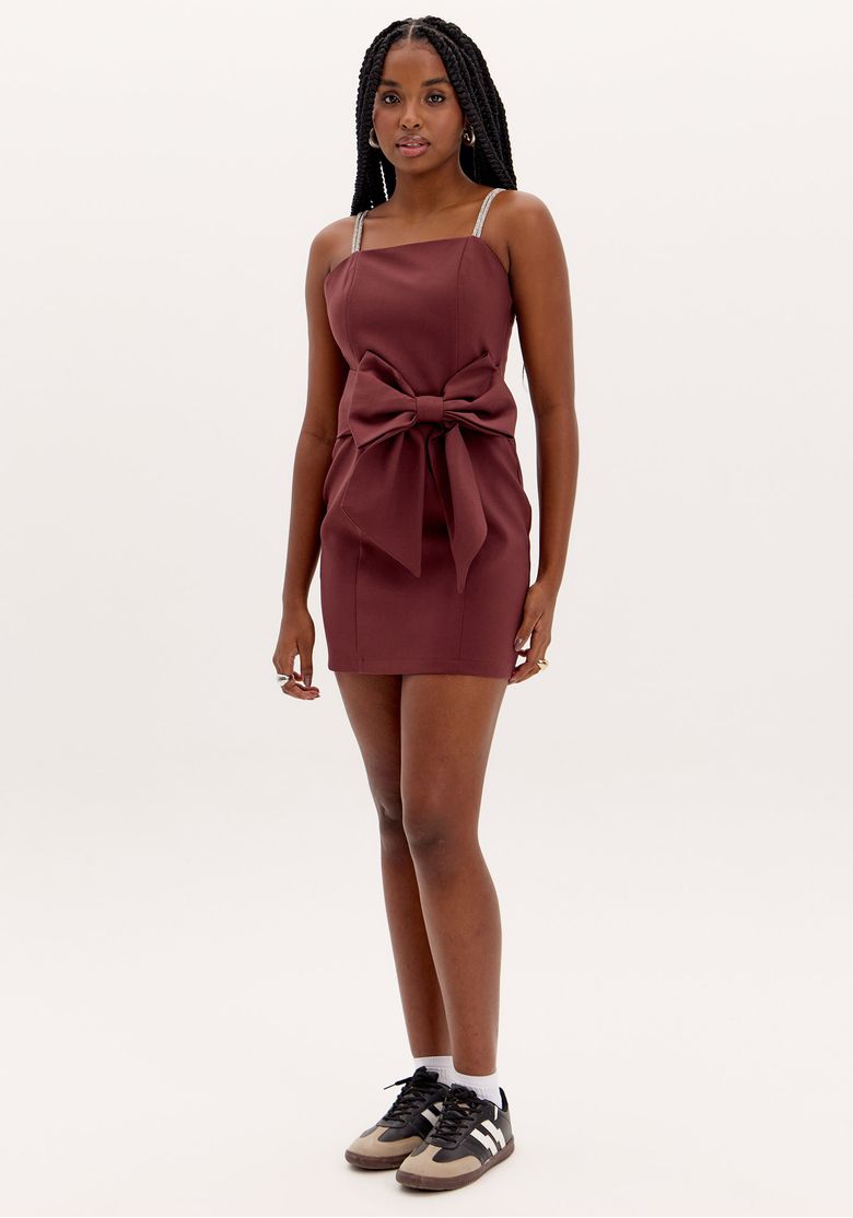 Modelo vestindo vestido curto marrom com maxi laço frontal, alças finas ajustáveis com brilho e caimento justo em plano de fundo neutro.