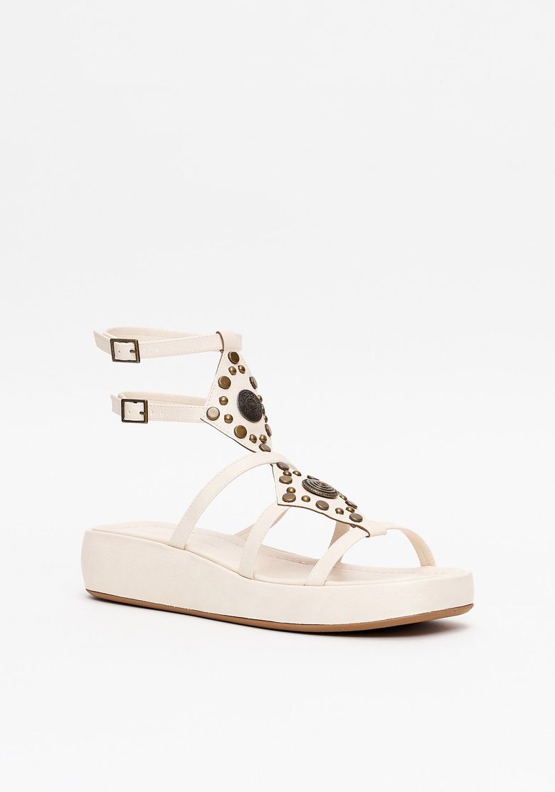 Close-up das tiras e aplicações boho com tachas e medalhão metálico na parte frontal da sandália flatform