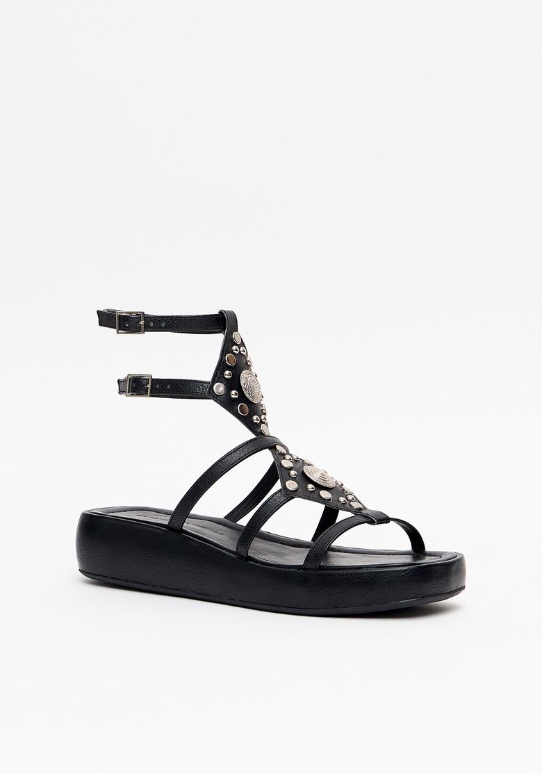 Close-up das tiras e aplicações boho com tachas e medalhão metálico na parte frontal da sandália flatform