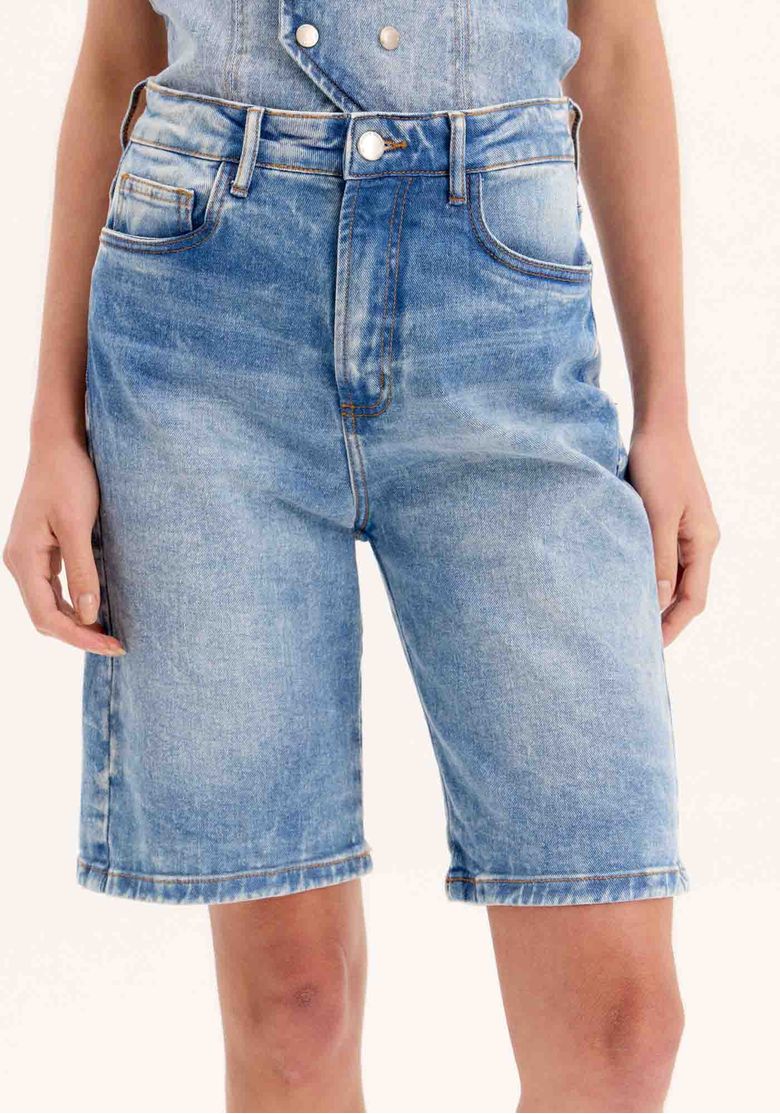 Close frontal da bermuda jeans alongada mostrando cós com passantes, fechamento por botão e zíper metálico e lavagem stone wash - detalhe bermuda jeans
