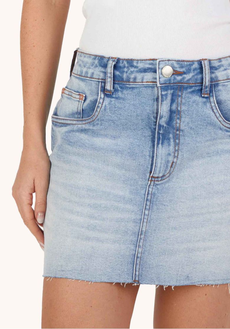 Modelo de costas usando saia curta jeans azul mostrando bolsos traseiros em formato de coração e barra desfiada