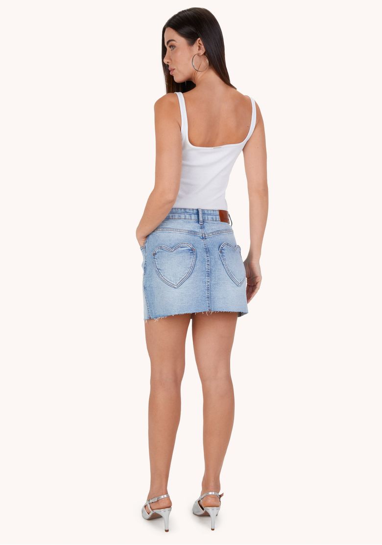 Close frontal da saia jeans destacando cós com passantes, botão metálico, costura central e barra levemente desfiada em denim azul claro