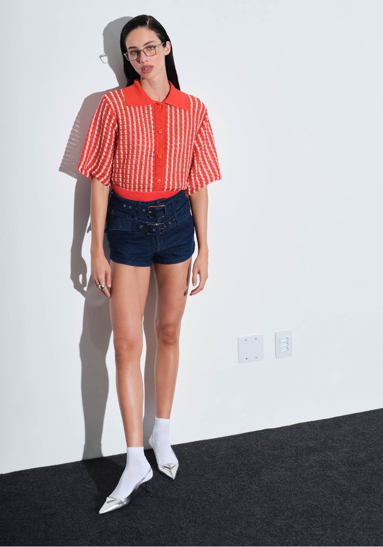 Modelo vestindo short jeans hot pants azul com cinto, combinado com blusa listrada e sapatos prateados.