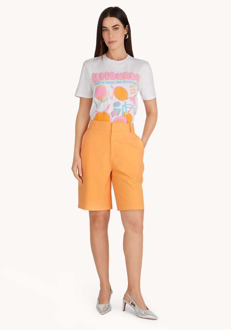 Modelo em pé vestindo bermuda de alfaiataria laranja frente completa com camiseta branca estampada, cintura alta e passantes