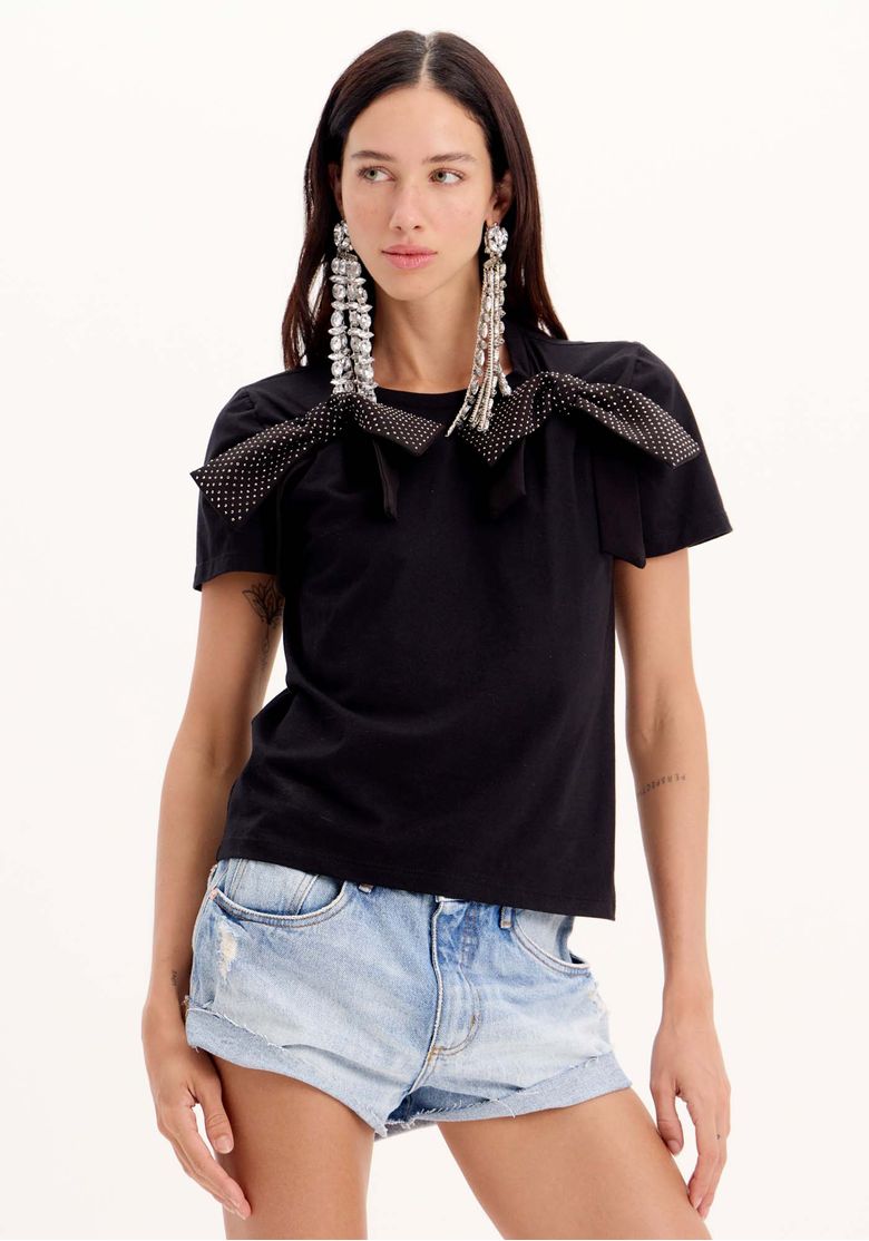 T-shirt feminina preta com manga curta e laços brilhantes, vista frontal em pose lateral com shorts jeans