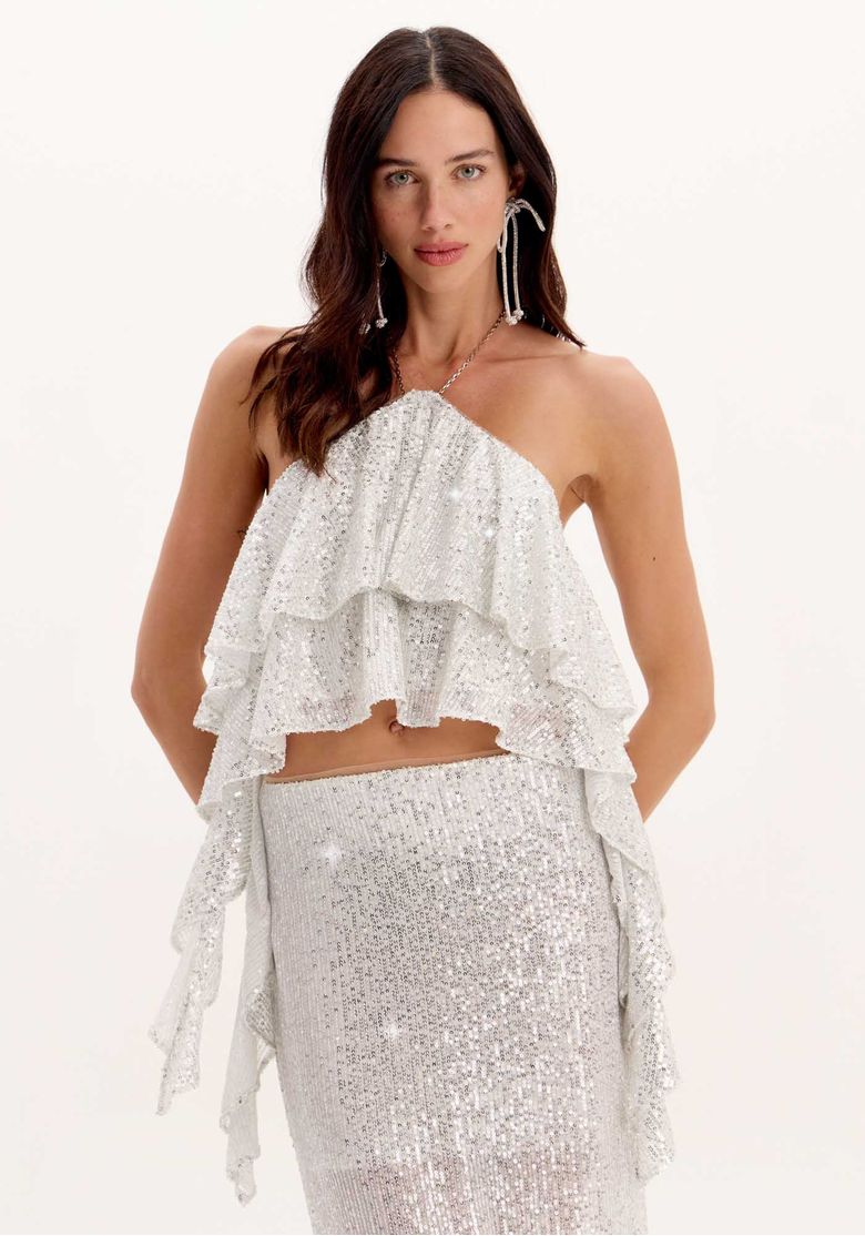 Modelo usando blusa cropped de paetês prateada com babados em camadas e alça halter fina, look festa com saia combinando