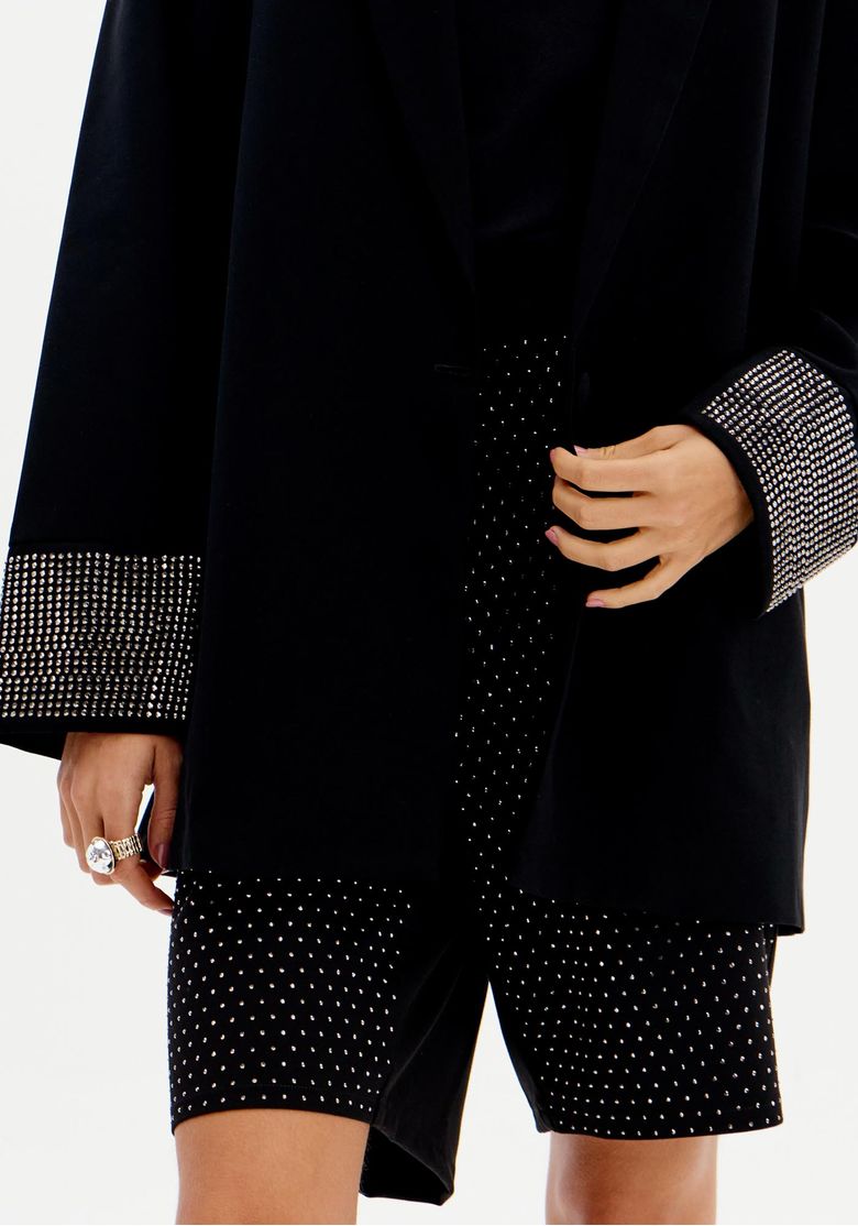 Close do blazer feminino preto oversized com mangas longas e detalhe brilhante nos punhos, em look sofisticado noturno