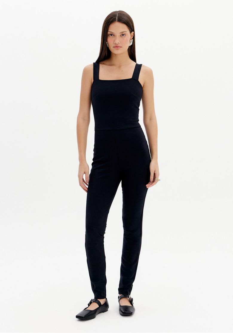 Modelo usando macacão longo preto feminino com alças largas, decote quadrado e modelagem ajustada em visão frontal com sapato preto