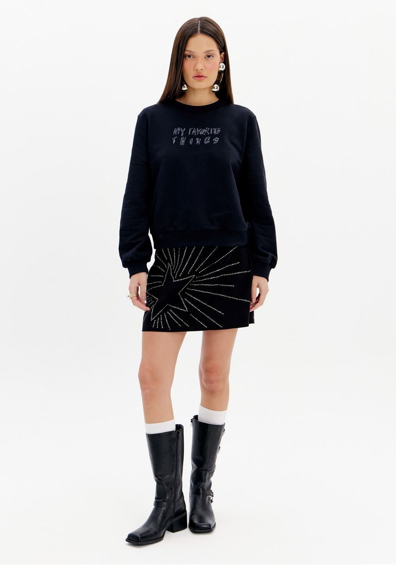 Modelo vestindo saia curta preta com aplicações de brilhos em formato de estrela, combinada com blusa preta e botas.