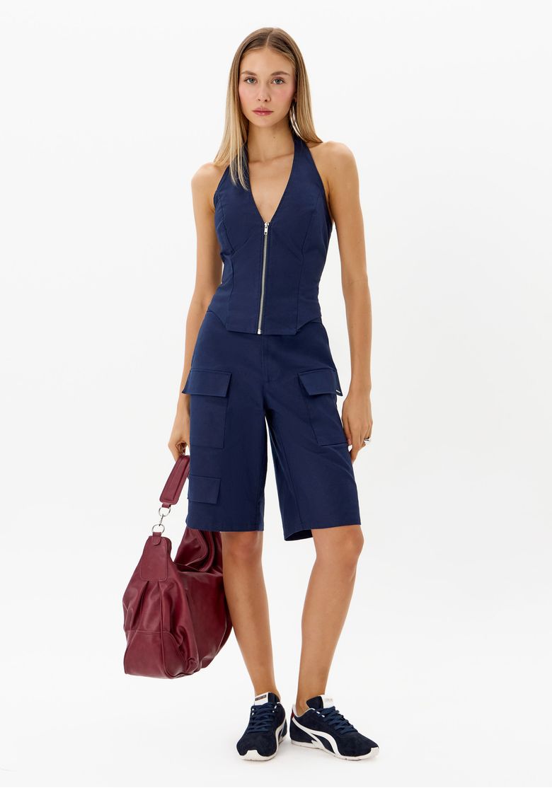 Look feminino com blusa frente única azul escuro e zíper frontal combinada com bermuda cargo azul, tênis escuro e bolsa vinho
