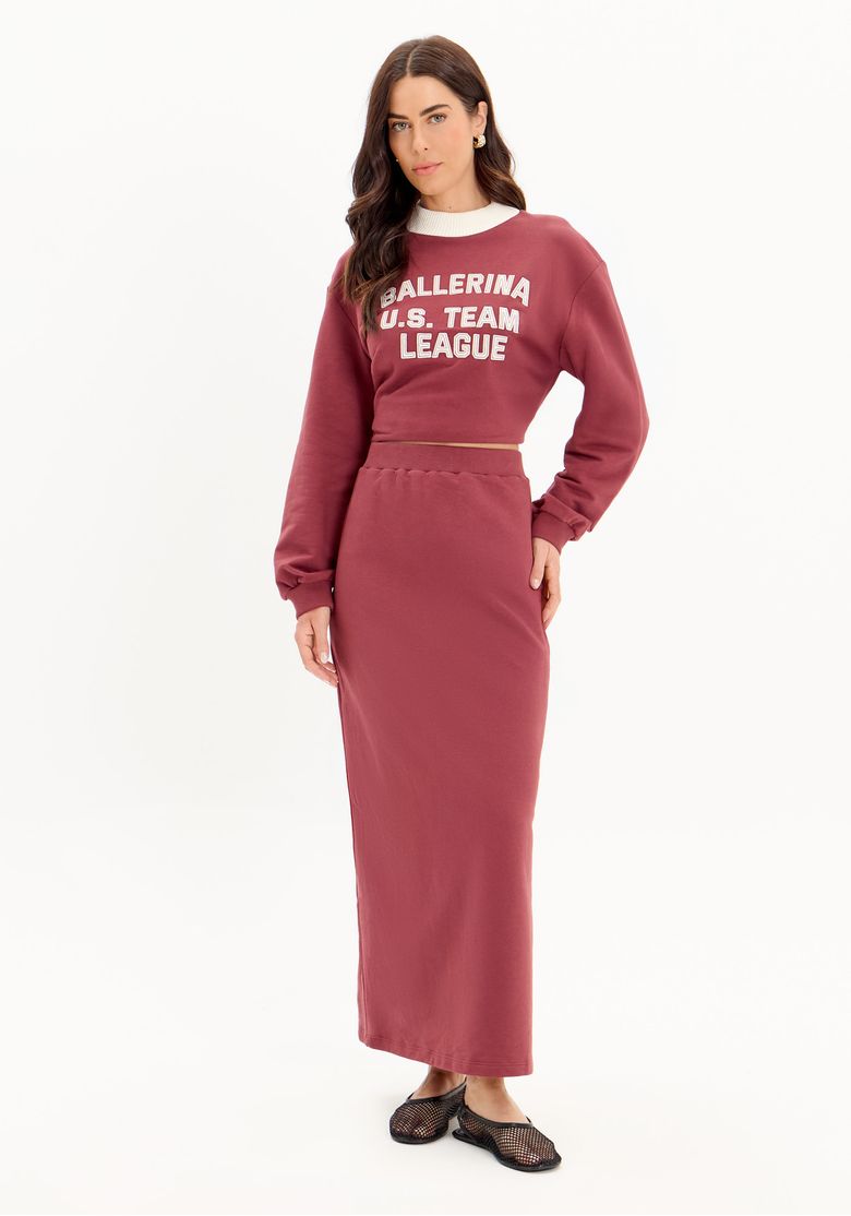 Modelo vestindo saia super midi de moletom vermelho com fenda traseira, combinada com top de moletom.