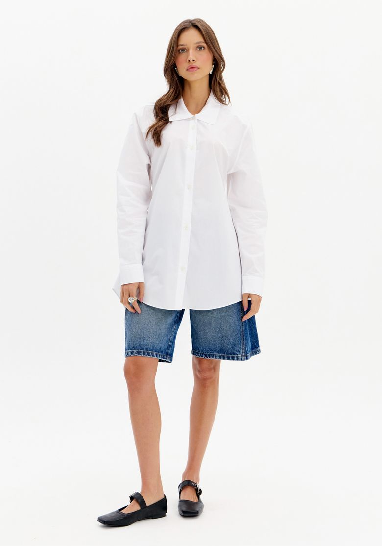 Modelo usando bermuda jeans feminina cintura alta azul com camisa branca oversized e sapato preto em look casual moderno