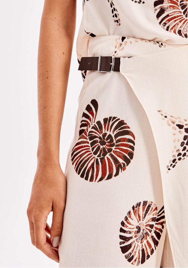 Close da calça reta estampada branca com detalhe de fivela lateral na cintura e sobreposição assimétrica, destacando a estampa tropical em tons terrosos