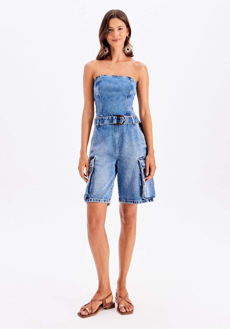 Modelo veste macacão curto jeans azul sem alças com cinto e bolsos utilitários laterais, em visão frontal completa