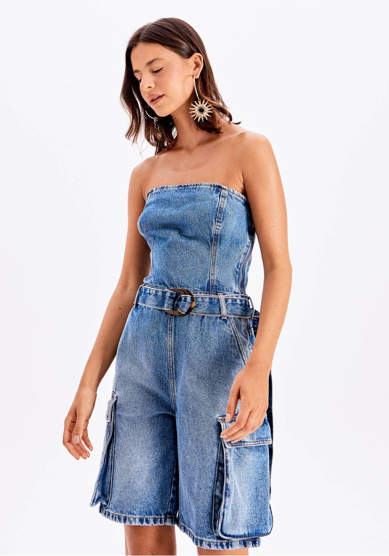 Modelo com macacão tomara que caia jeans azul curto, cintura marcada por cinto e pernas amplas de comprimento acima do joelho