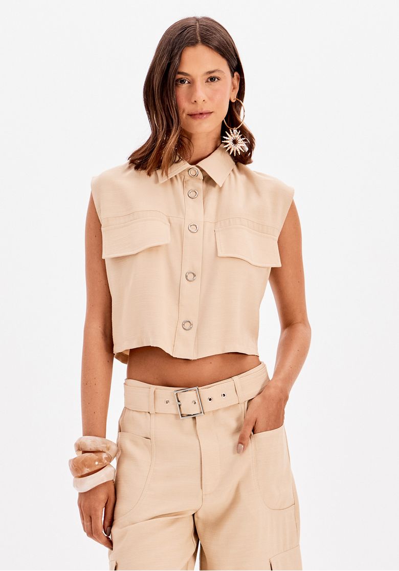 Modelo vestindo camisa cropped sem mangas bege vista frontal com gola clássica e bolsos de lapela, tecido plano leve