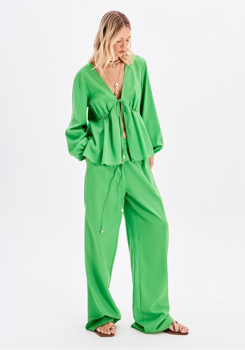 Modelo em pé usando calça full length verde em linho com cós elástico e blusa amarrada, vista frontal 3/4 - calça linho verde