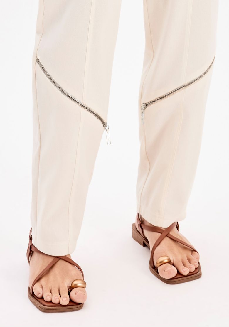 Close da calça balloon branca feminina com detalhe de zíper diagonal na barra e sandália rasteira marrom