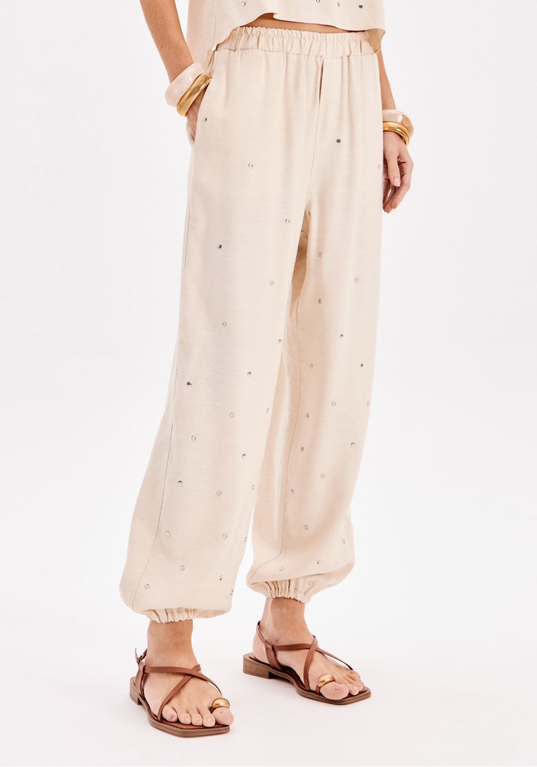 Close frontal de calça jogger bege com aplicações discretas, cós elástico e barra com punho, detalhe de modelagem ampla feminina