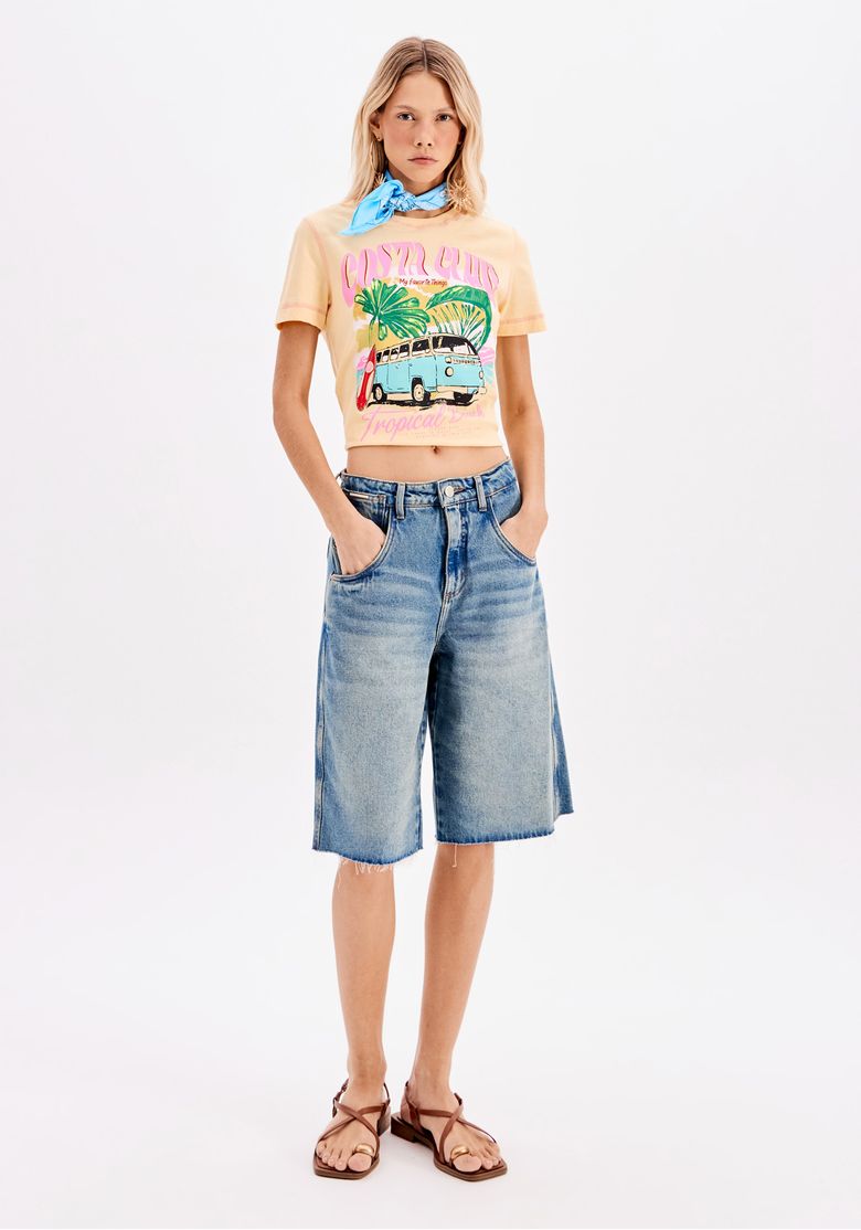 Modelo em pé vestindo camiseta baby look amarela estampada com tema tropical, combinada com bermuda jeans e sandálias, formando um visual casual urbano em estúdio.