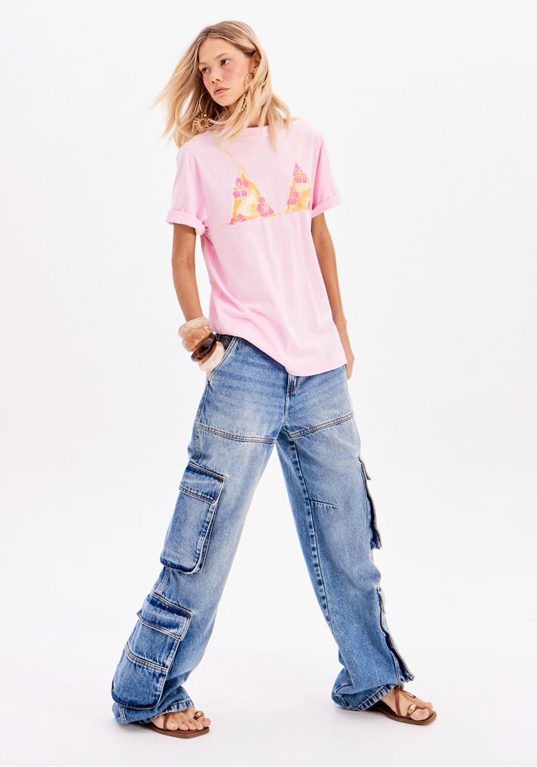 Modelo usando camiseta oversized rosa com estampa de biquíni, combinada com calça jeans cargo.