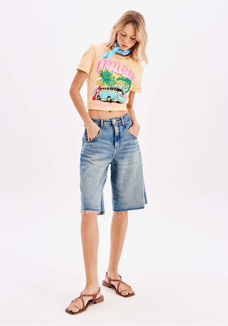 Close frontal destacando a estampa da camiseta amarela, composta por uma Kombi azul, folhas tropicais e tipografia rosa, junto com um lenço azul no pescoço.