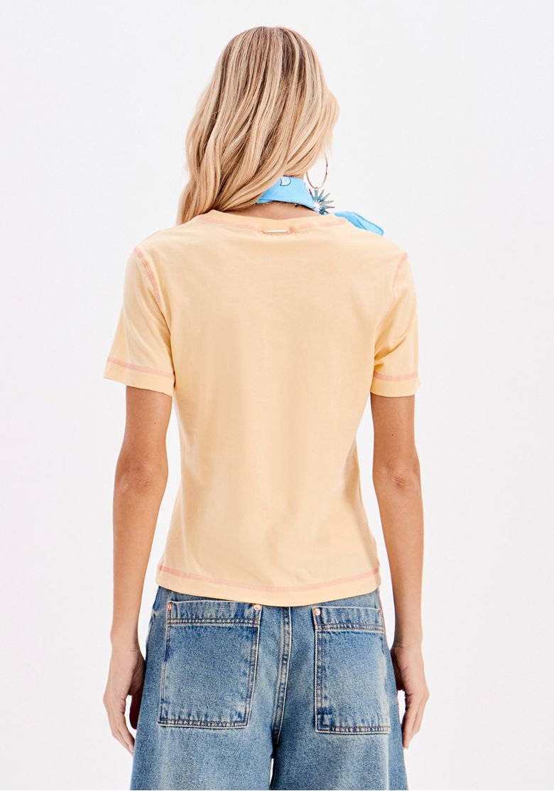 Verso da camiseta baby look amarela apresentando barra reta e costas lisas, combinada com bermuda jeans de bolsos traseiros.