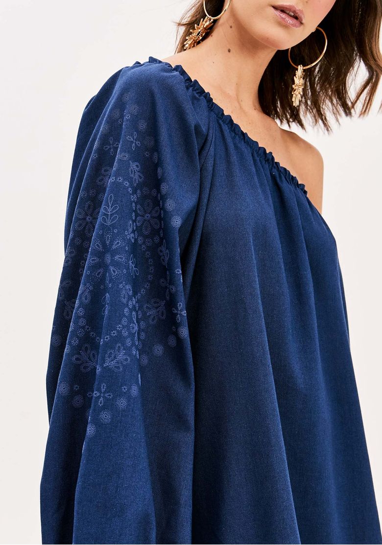 Modelo usando vestido ombro único azul vista três quartos, mostrando caimento fluido e manga bufante com detalhe bordado