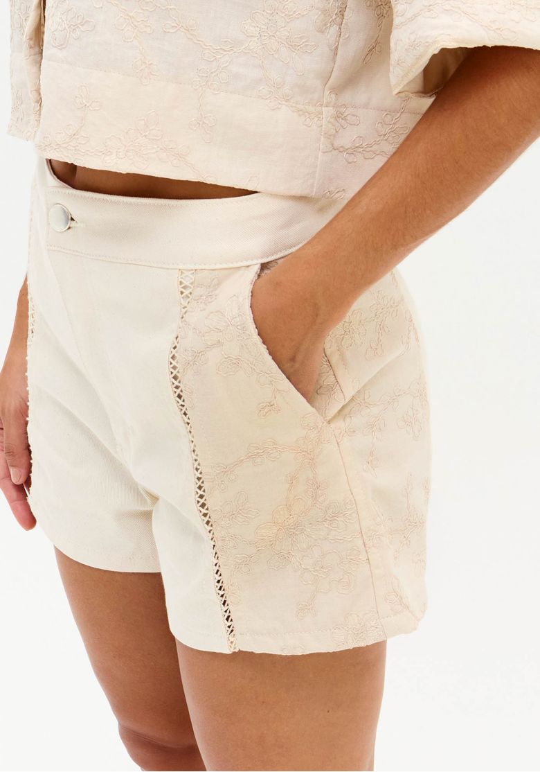 Close do short branco mostrando cós alto com botão, bolso lateral e recorte com detalhe vazado em sarja