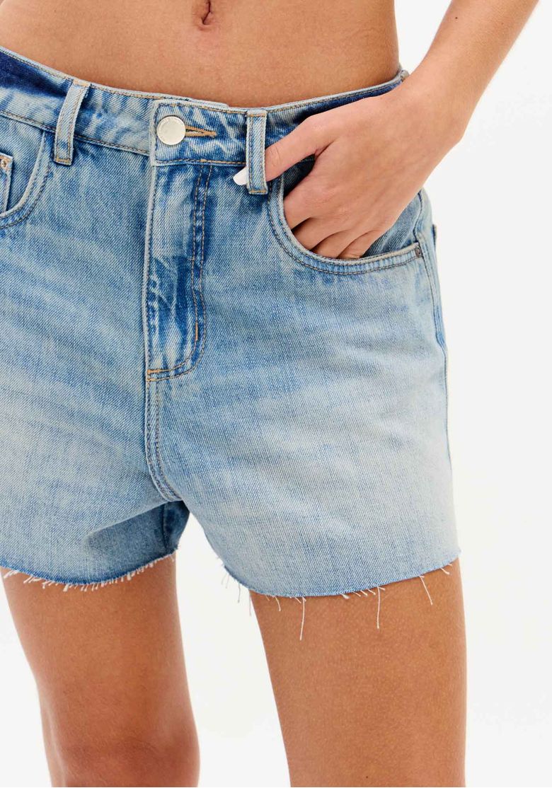 Close do cós e bolso frontal do short jeans feminino cintura alta azul claro com lavagem clara e barra desfiada