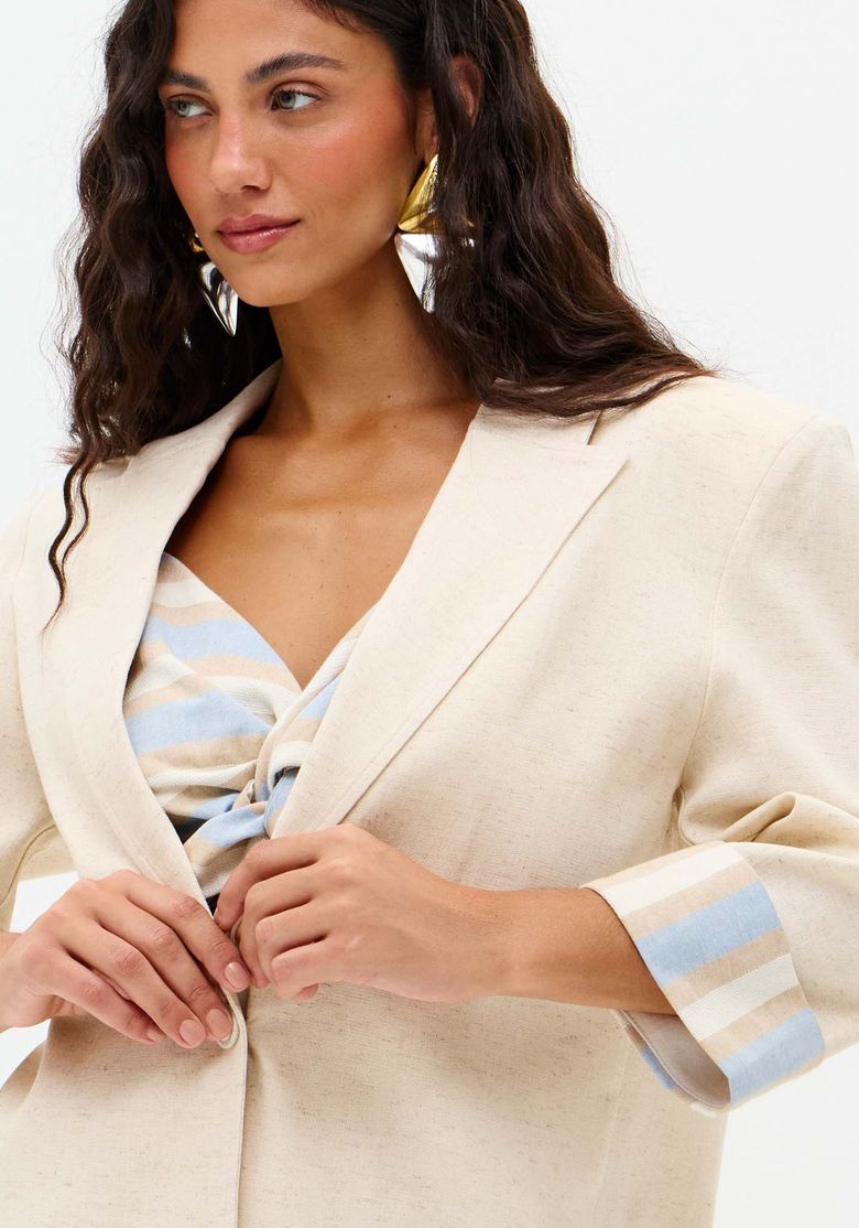 Detalhe frontal do blazer alongado bege com fechamento por um botão forrado e lapela clássica, mostrando o forro listrado nos punhos - detalhe blazer linho
