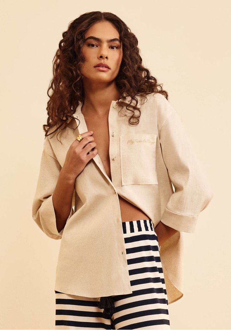 Modelo com camisa feminina bege oversized aberta, manga 7/8 e bolso frontal, combinada com calça listrada