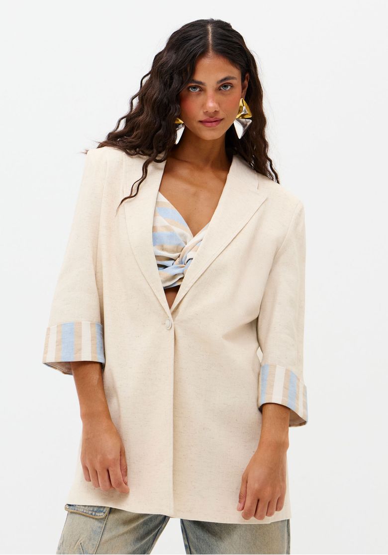 Vista frontal do blazer alongado bege fechado, modelagem tradicional e caimento descolado, mangas 3/4 com punhos listrados expostos - terceira peça elegante