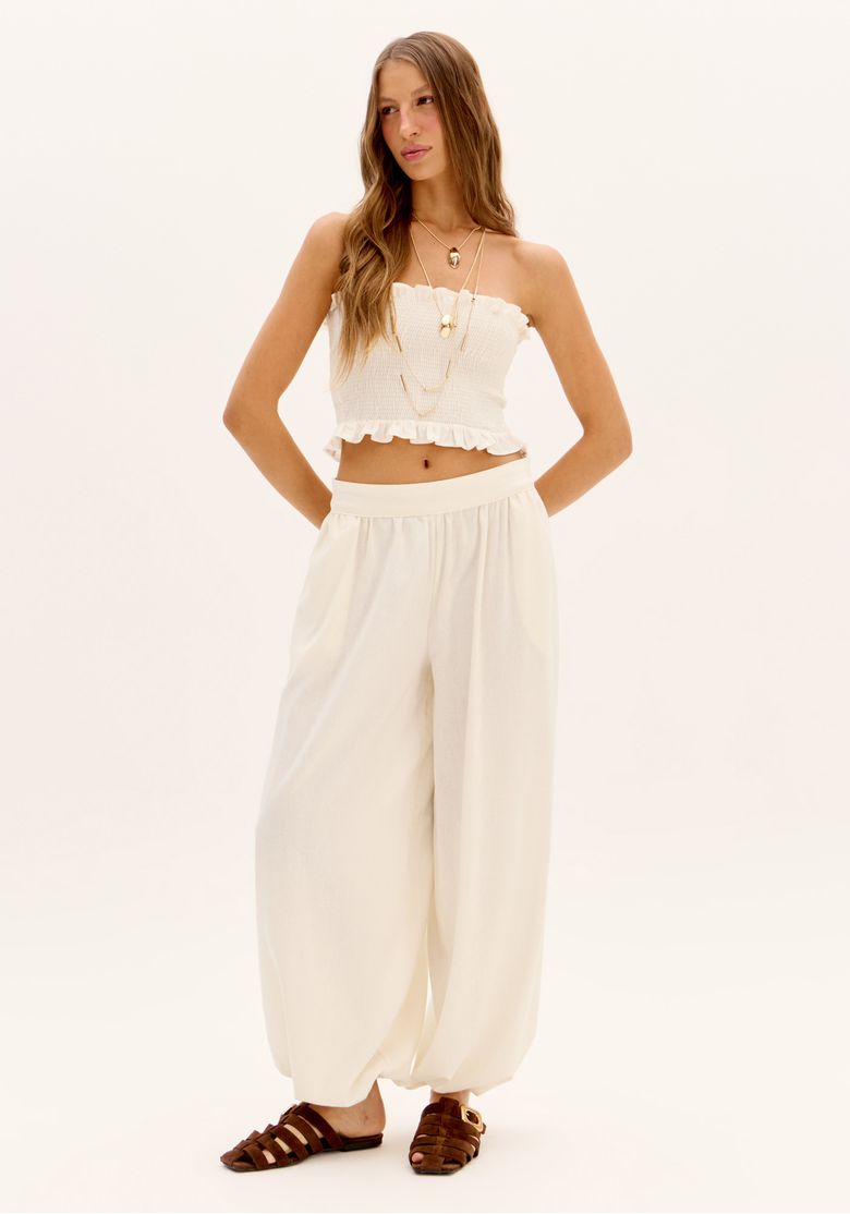 Modelo em corpo inteiro com top cropped branco sem alça e calça pantalona clara, visual leve para sunset
