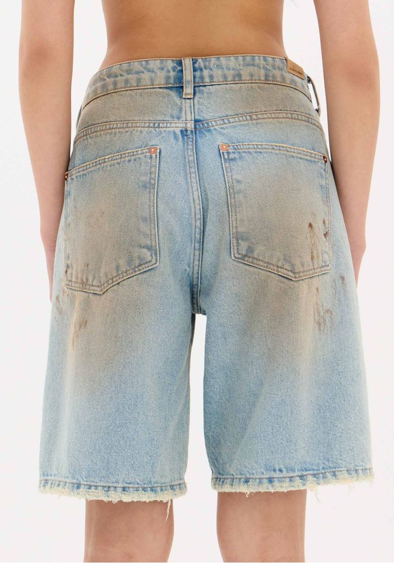 Vista traseira da modelo com bermuda jeans de cintura média azul estonada, jaqueta de couro marrom e botas, mostrando o comprimento até acima dos joelhos