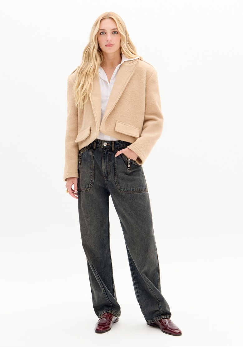 Modelo vestindo calça jeans com modelagem baggy, cintura média e lavagem escura, combinado com blazer bege e sapatos vinho - vista frontal corpo inteiro.