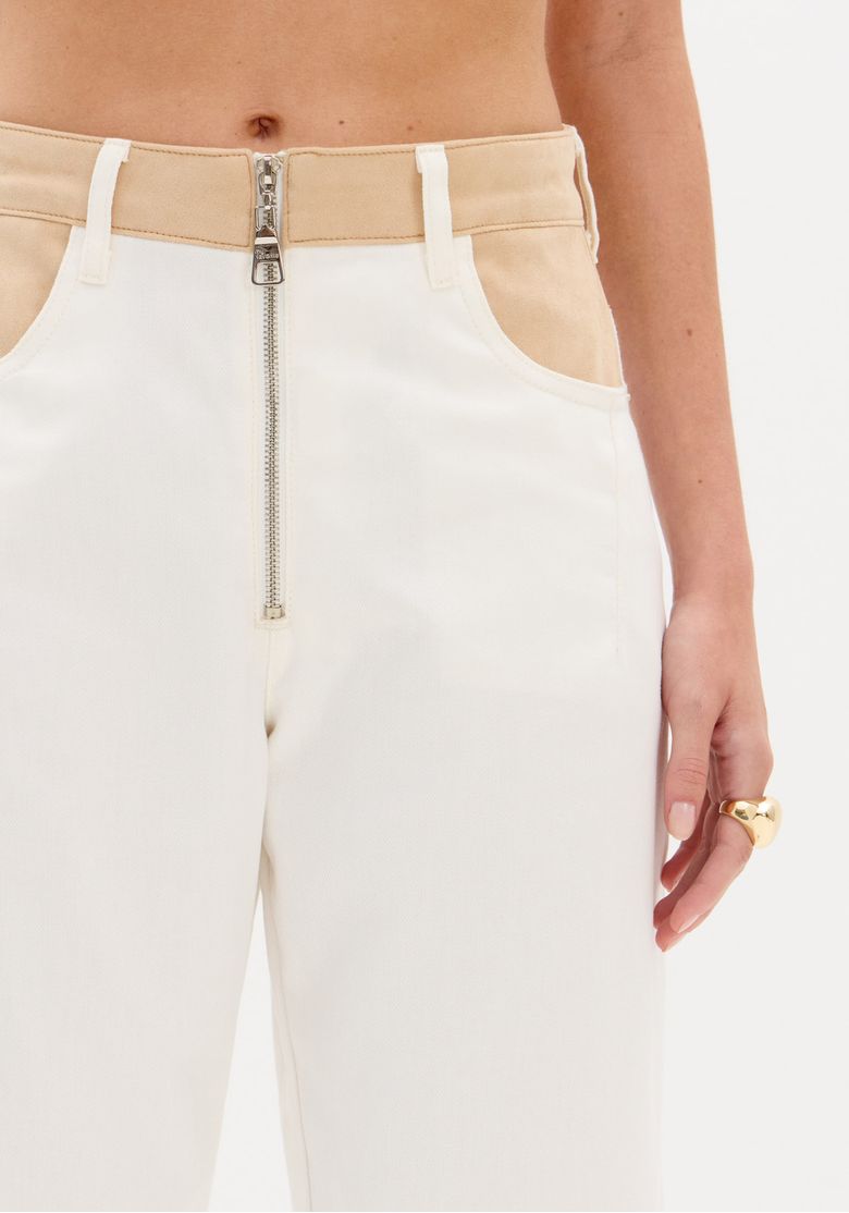 Close no cós em contraste bege da calça em sarja, apresentando passantes, zíper frontal exposto e detalhe do bolso frontal.