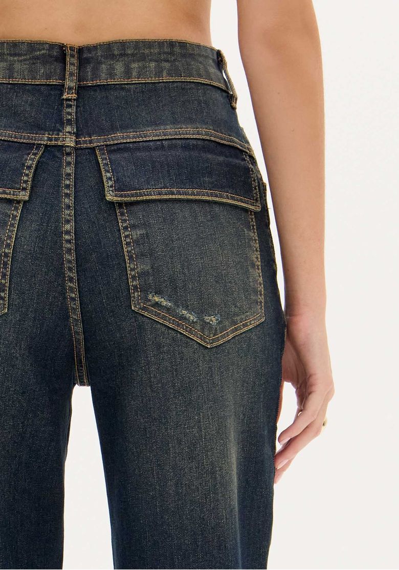 Detalhe do bolso traseiro com aba na calça jeans lavagem escura, costura e leve desgaste do denim