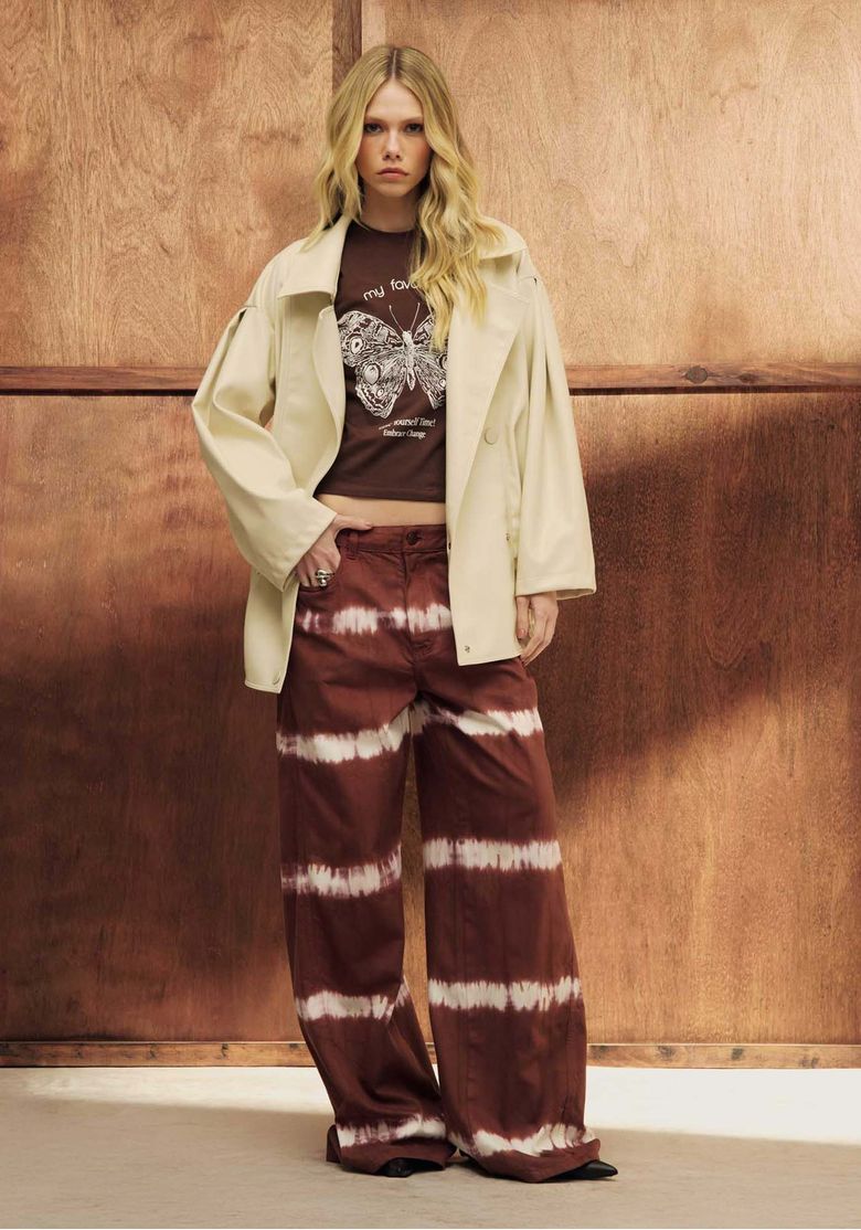 Modelo usando calça sarja wide leg tie dye marrom com faixas horizontais brancas em estúdio com painel de madeira, cintura média e cós com passantes