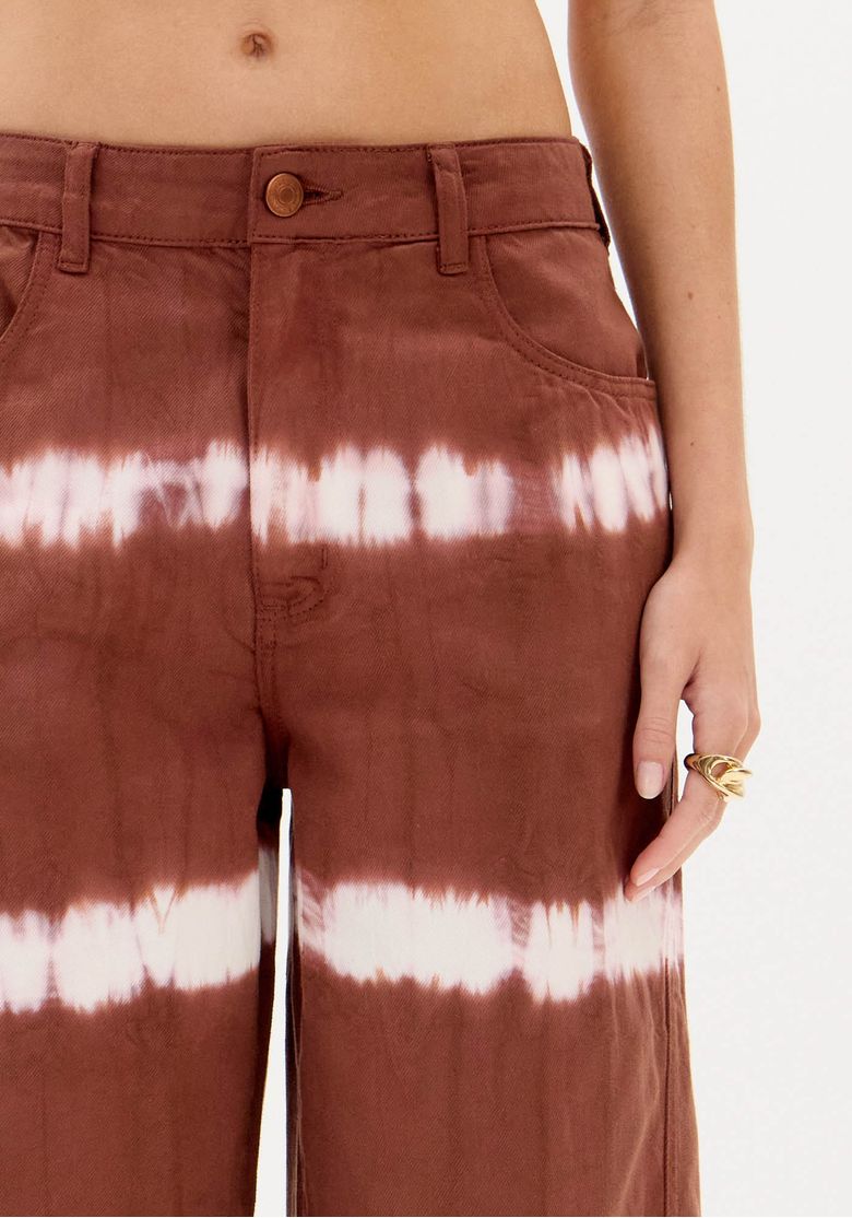 Close frontal da calça sarja marrom tie dye mostrando cós com passantes, botão metálico e bolsos frontais