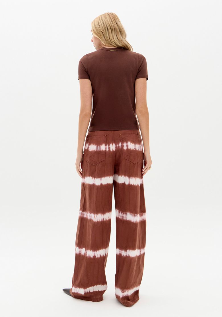 Vista traseira da modelo com camiseta marrom e calça sarja wide leg tie dye, destacando bolsos traseiros chapados e padrão de faixas