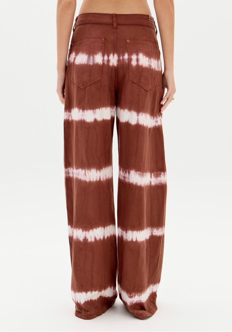 Vista traseira da calça sarja tie dye marrom em fundo branco, evidenciando o comprimento longo e o caimento da perna