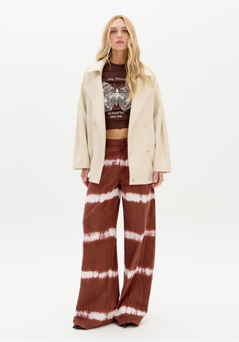 Modelo em estúdio branco usando jaqueta creme e calça sarja wide leg tie dye marrom, look com silhueta fluida e atitude
