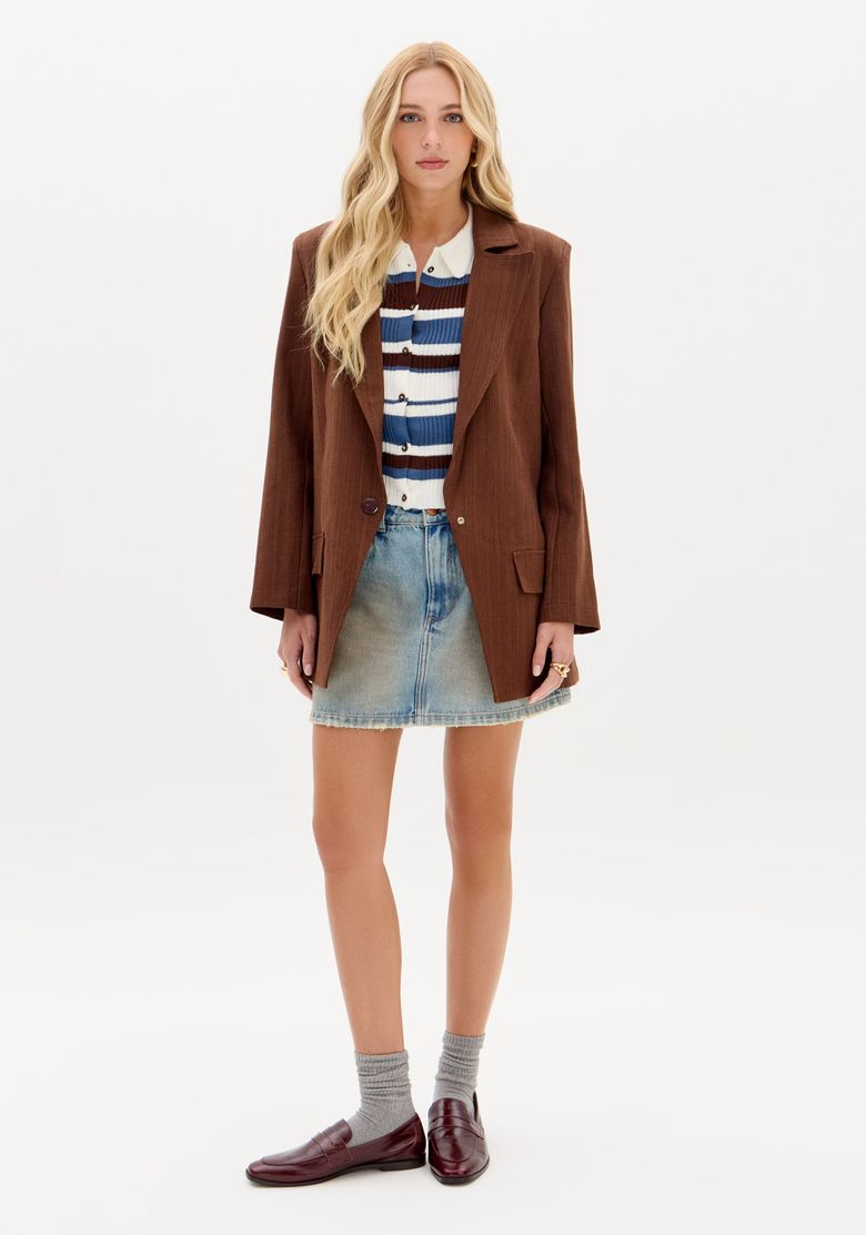 Modelo em pé usando saia jeans curta cintura média em denim azul com lavagem vintage, combinada com blazer marrom e top listrado; look completo com meias e mocassim.