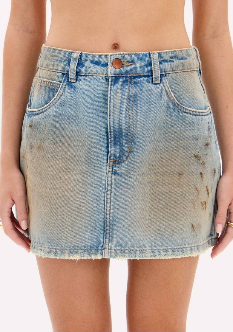 Detalhe frontal da saia jeans curta mostrando cós com passantes, botão, zíper e acabamento com barra desfiada em lavagem dirty.