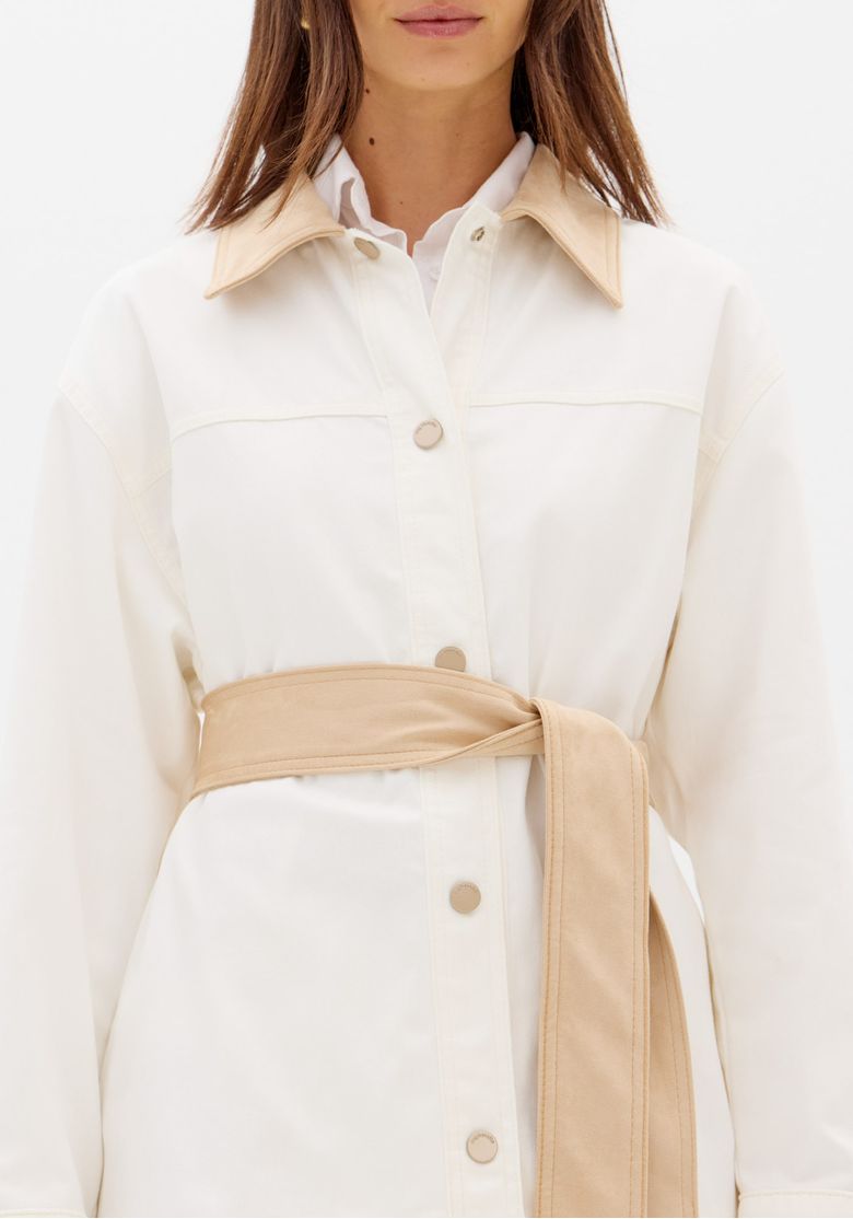 Modelo veste parka alongada branca bicolor com faixa na cintura, shorts off-white e botas marrom escuro, look completo