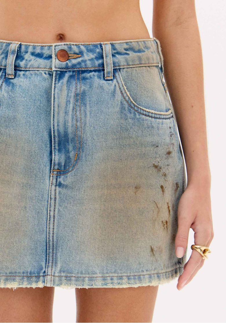 Close no bolso traseiro e costura da saia jeans curta, destacando a lavagem vintage e o efeito desgastado.