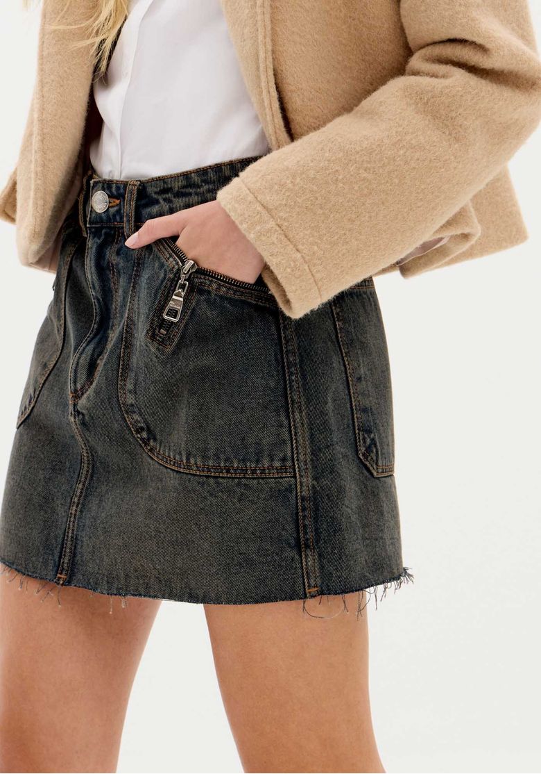Imagem lateral da saia jeans curta com mão no bolso, costura central e acabamento da barra desfiada em tecido 100% algodão.